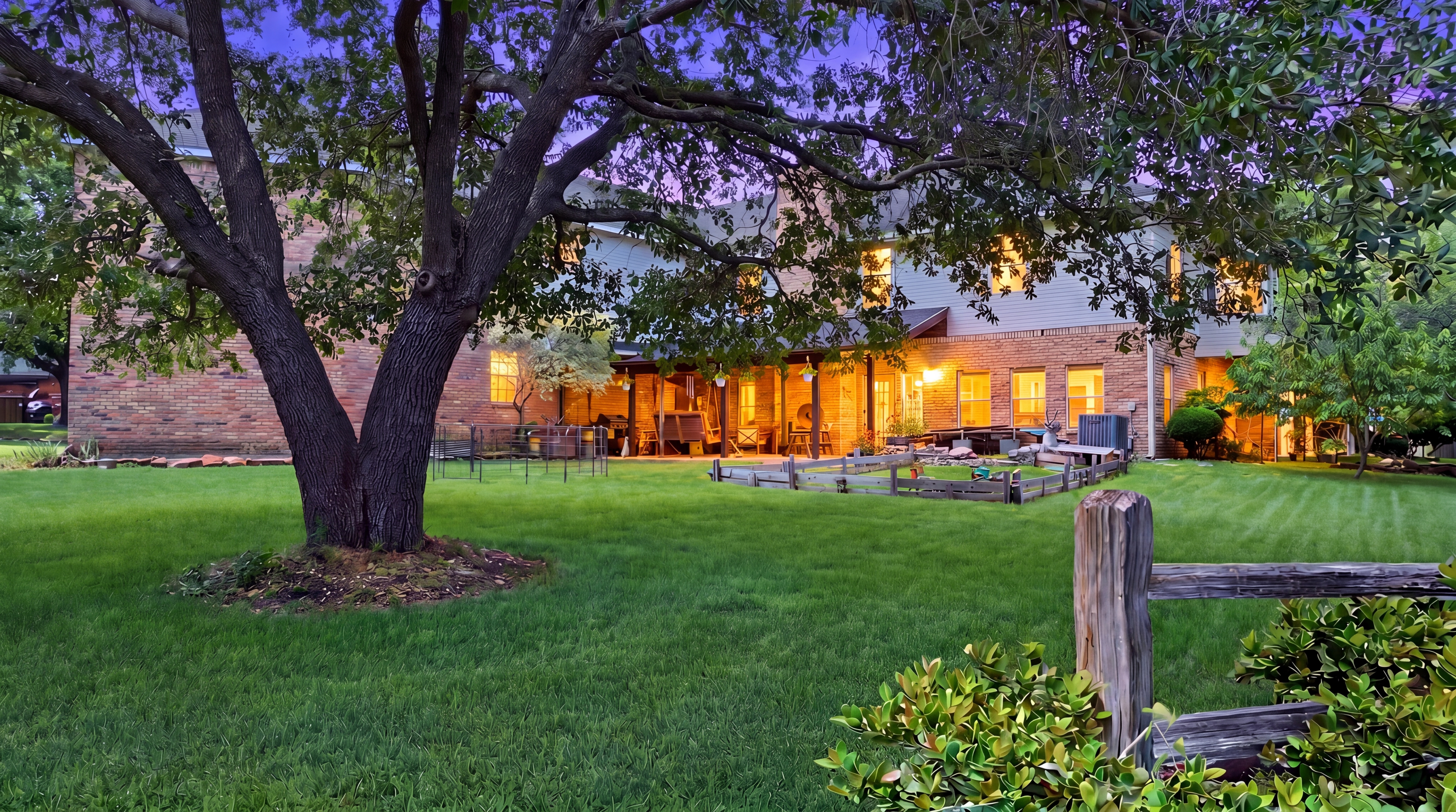 Rear yard at dusk with the home's brick wing illuminated, viewed past a large oak tree.